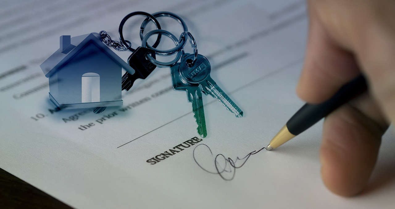 Une main signant un contrat, avec les clés d'un maison à côté.